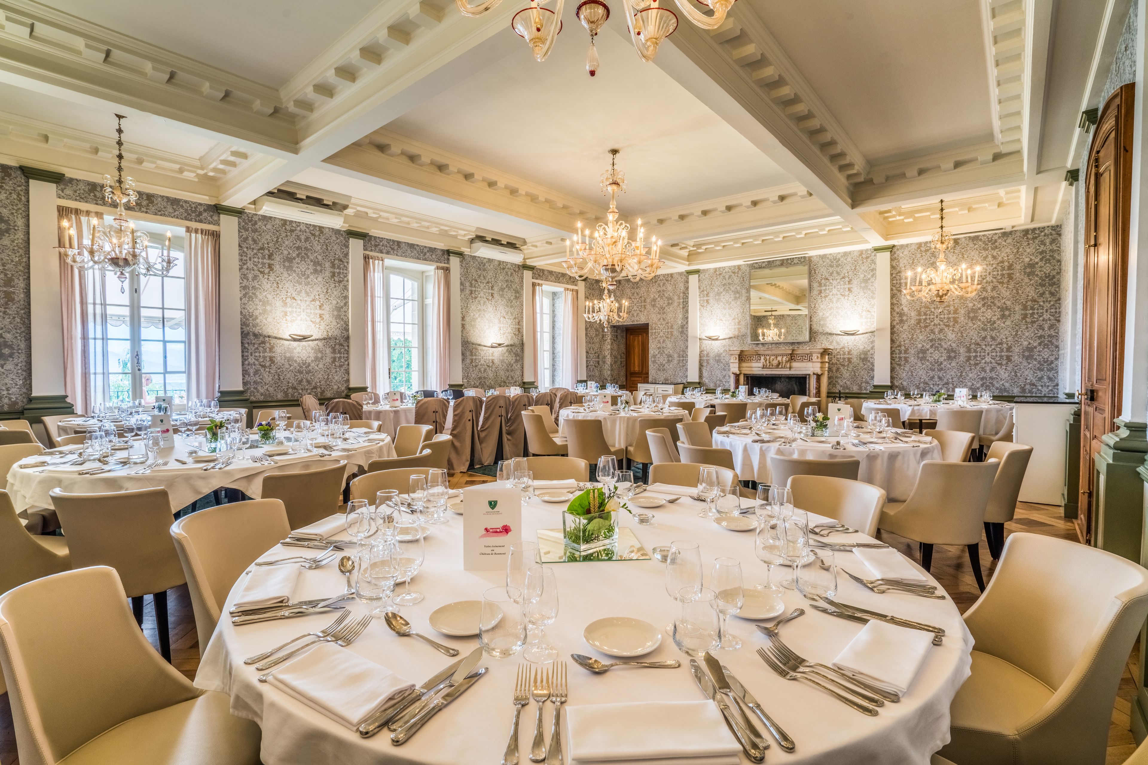 Une vaste salle de banquet avec des tables rondes dressées pour le repas, des lustres et une cheminée.