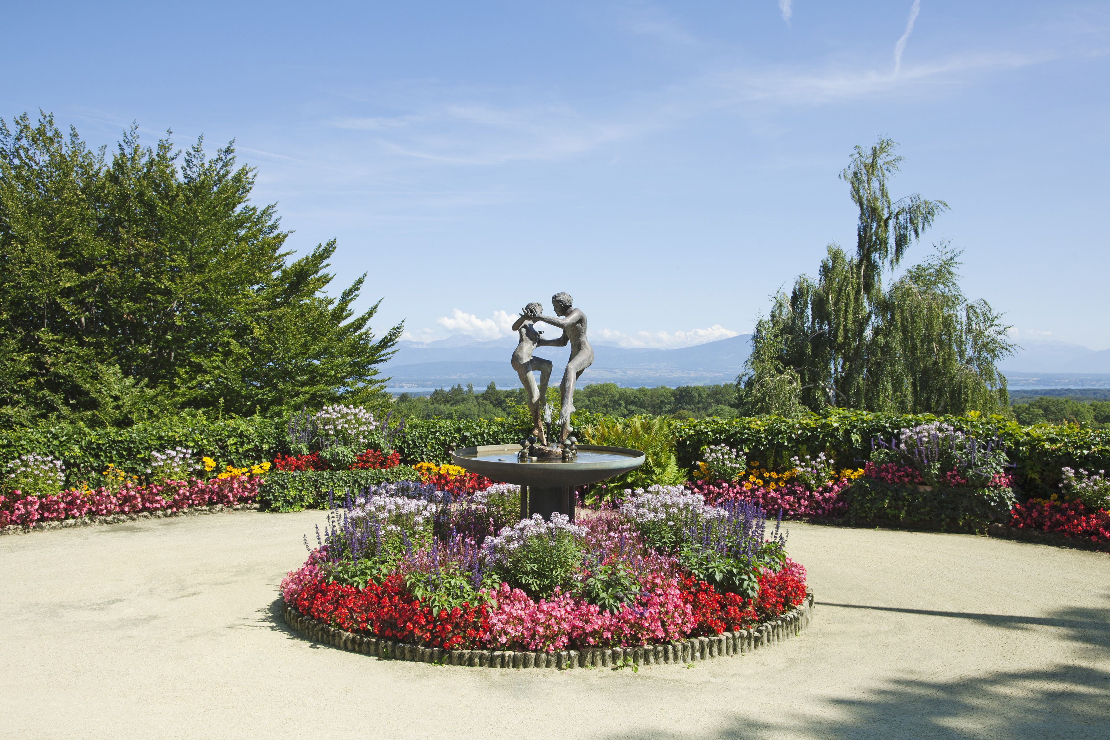 A garden with a central fountain and statues, surrounded by colorful flower beds and trees.