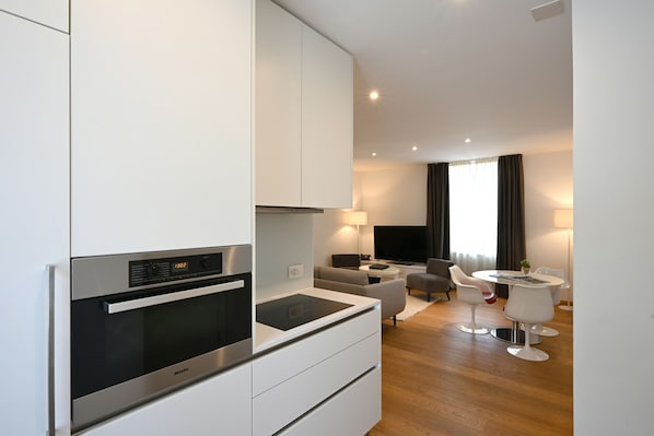 Una cocina moderna con electrodomésticos integrados, un comedor con una mesa redonda y sillas blancas, y una sala de estar con un sofá y una televisión.