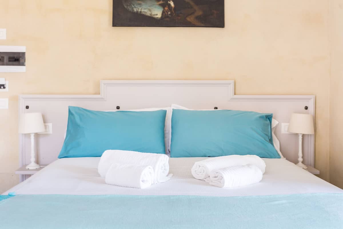 Una cama bien hecha con dos almohadas, dos toallas enrolladas y dos lámparas de noche.