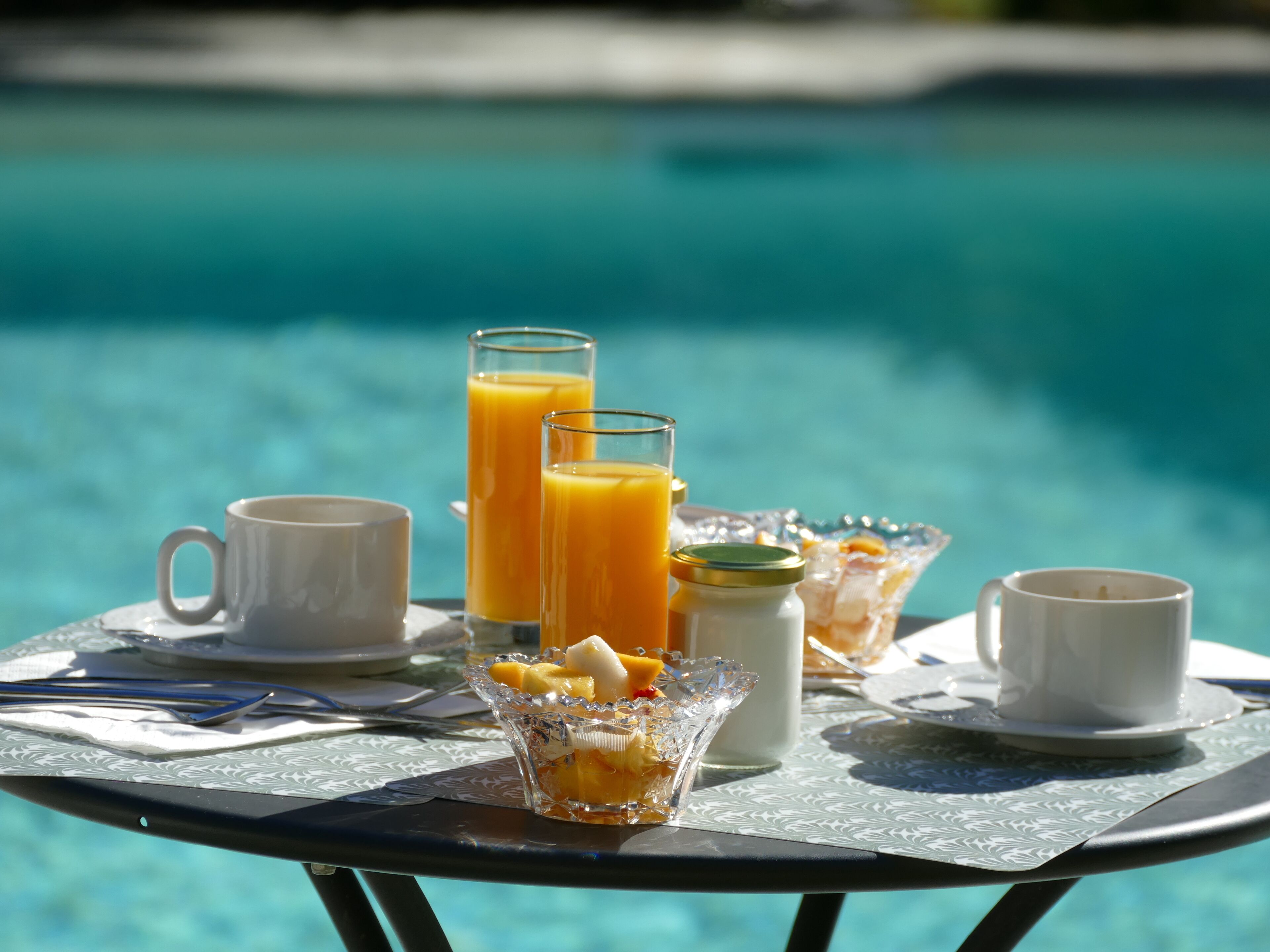 A breakfast setup by a pool with orange juice, milk, and a fruit bowl.