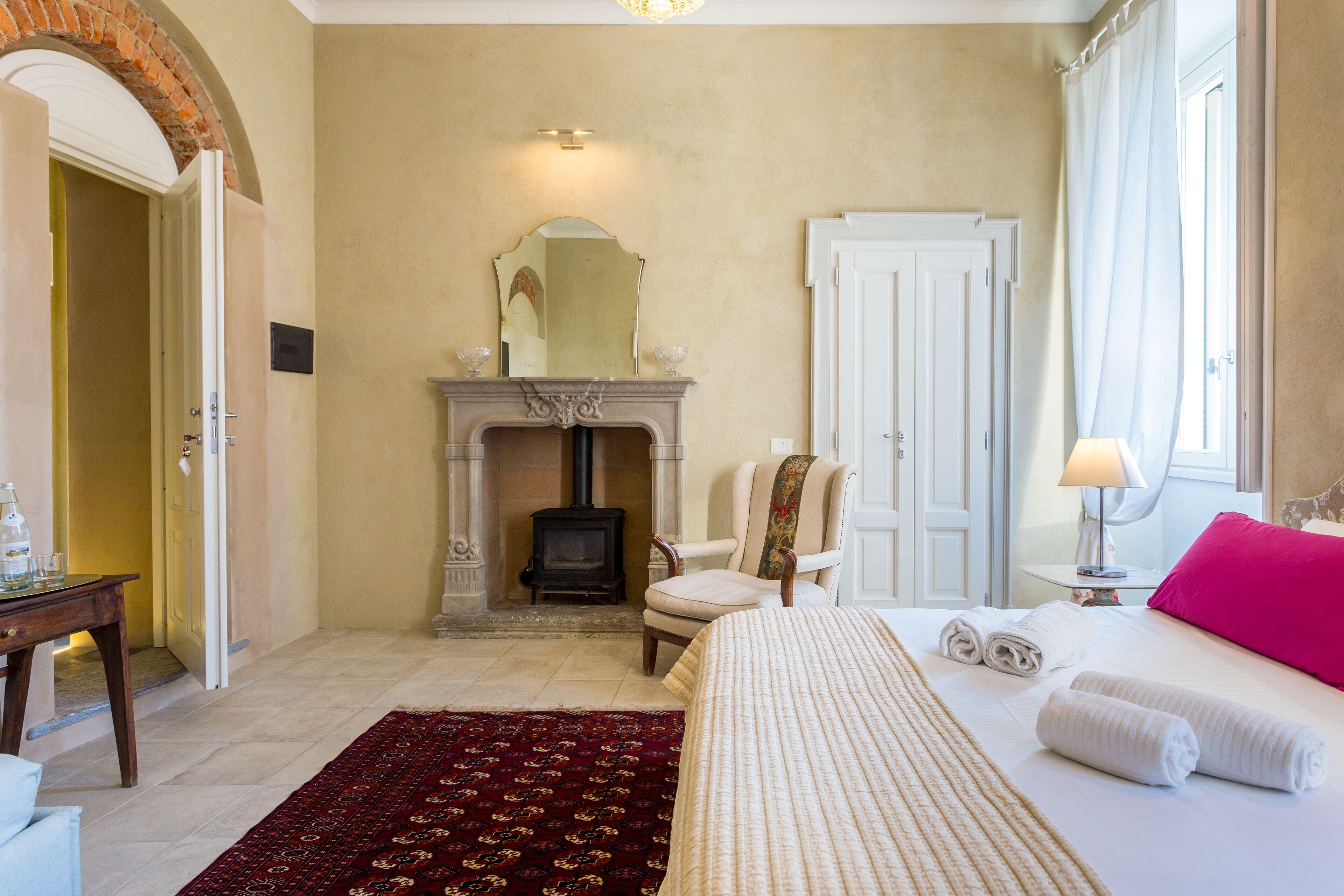 A hotel room with a fireplace, a bed with rolled towels, a chair, and a table with a lamp.