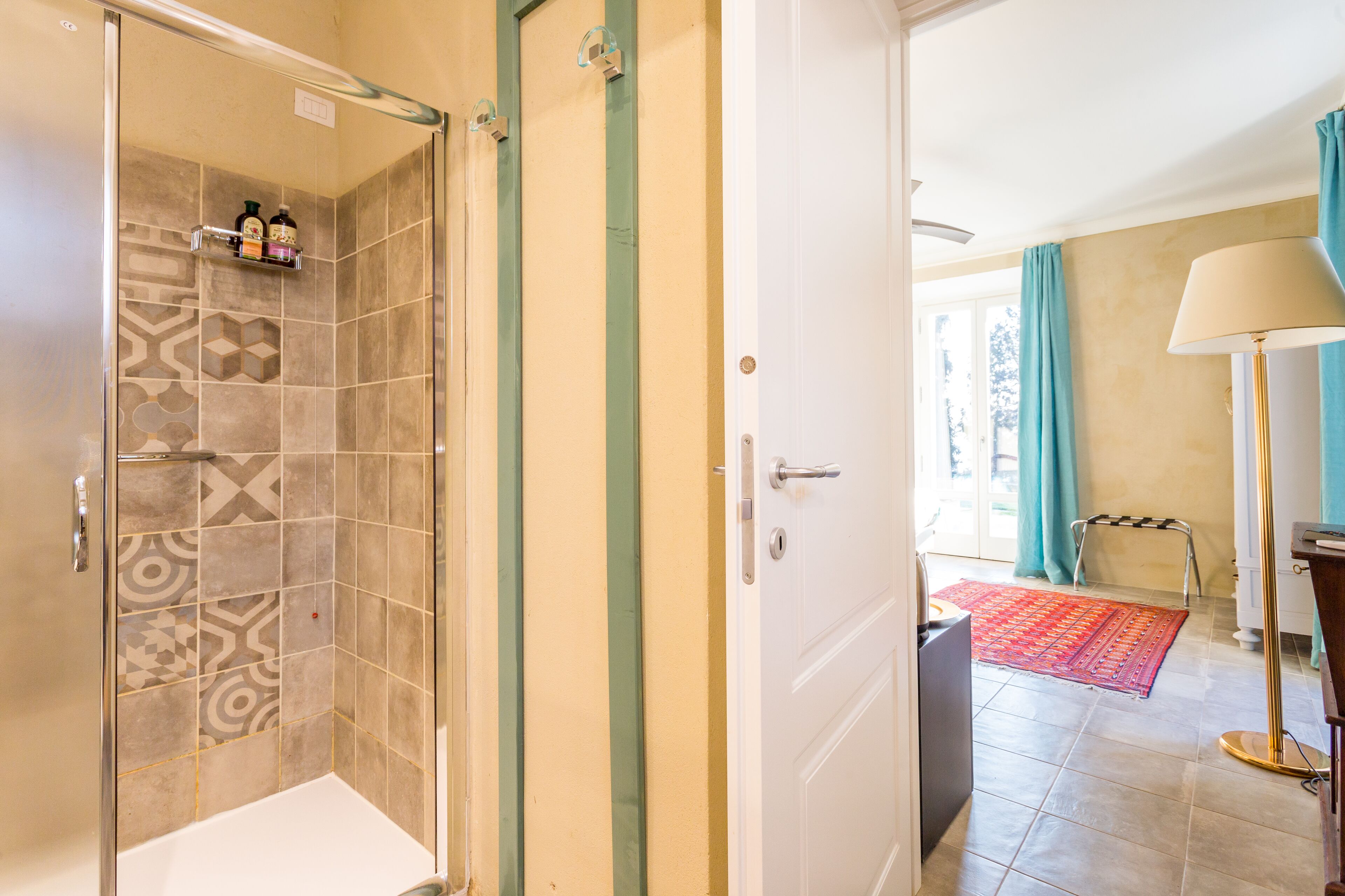 Une salle de bain avec des carreaux décorés, une douche et une porte donnant sur une pièce où se trouvent un tapis et une lampe.
