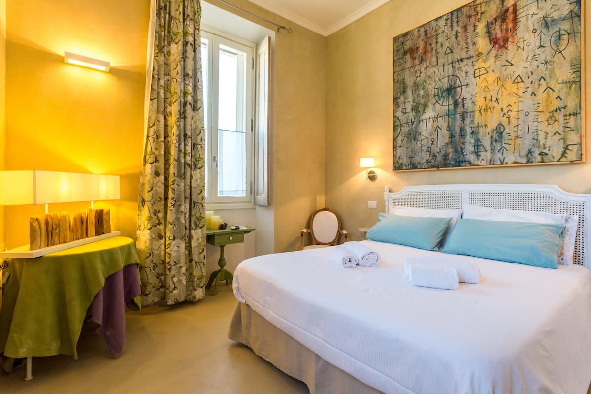 Una habitación de hotel con una cama, una mesita de noche con lámpara, un espejo, un cuadro en la pared y una ventana con cortinas.