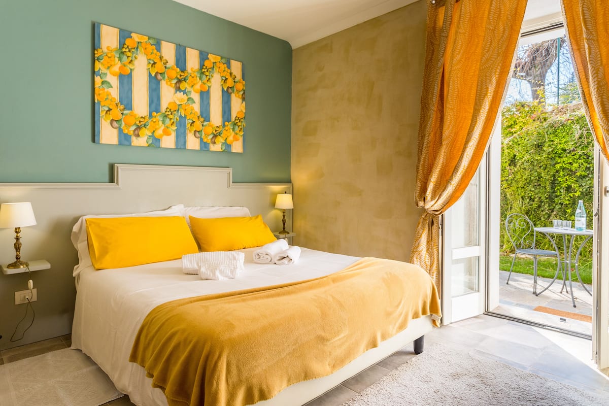 Habitación de hotel con cama, mesita de noche con lámpara, ventana con cortinas y vistas a zonas verdes.