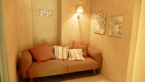 Un rincón acogedor con un sofá color crema, dos almohadas, una lámpara de pared y dos cuadros con motivos florales.
