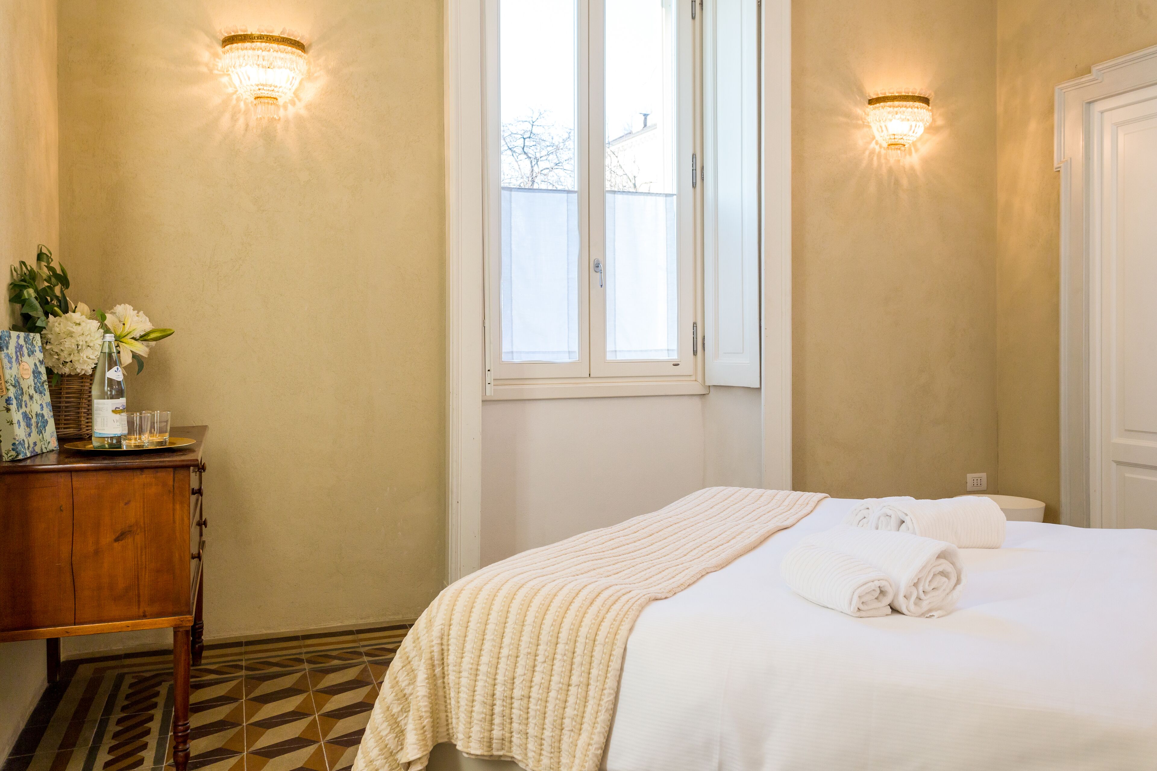Una habitación de hotel con una cama, una mesita de noche de madera, una ventana y dos apliques de pared.