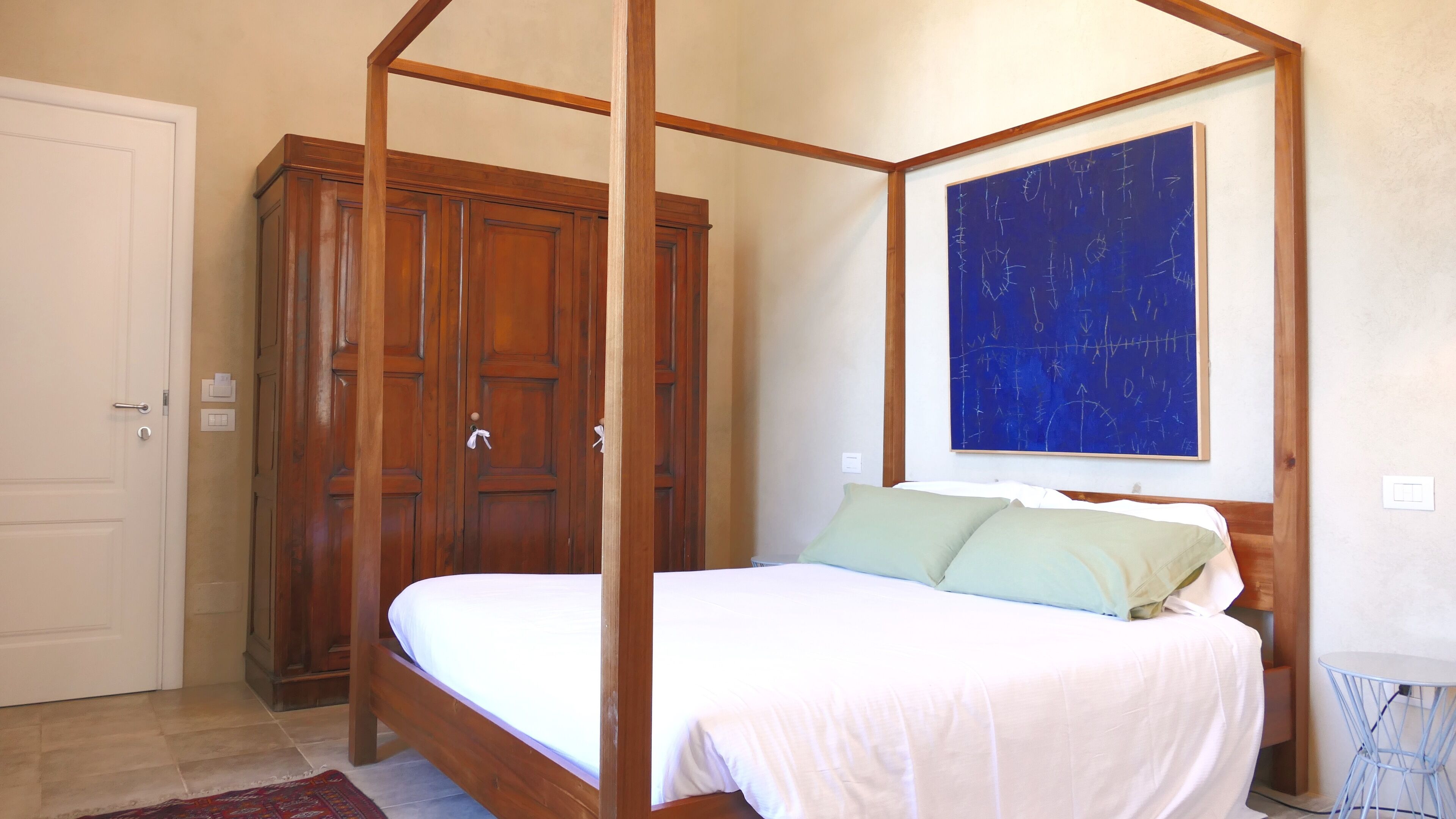 A bedroom with a four-poster bed, wooden wardrobe, and a blue abstract painting on the wall.