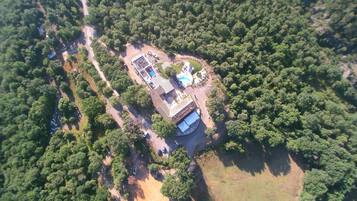An aerial view of a property with a swimming pool, a large building, and surrounding forested area.