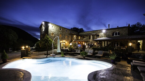 Una piscina con borde de piedra, mobiliario exterior y un edificio con múltiples ventanas e iluminación.