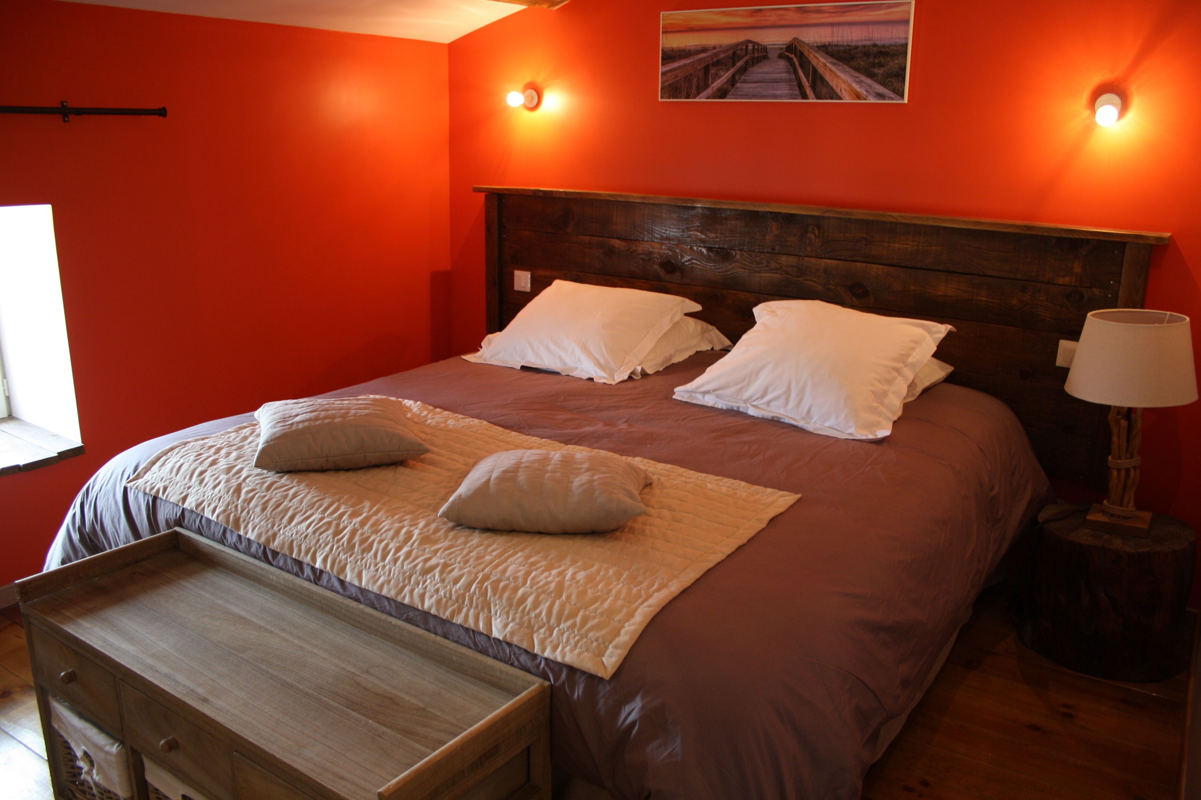 Un dormitorio con cabecera de madera, dos almohadas blancas, un banco de madera y una mesita de noche con una lámpara.
