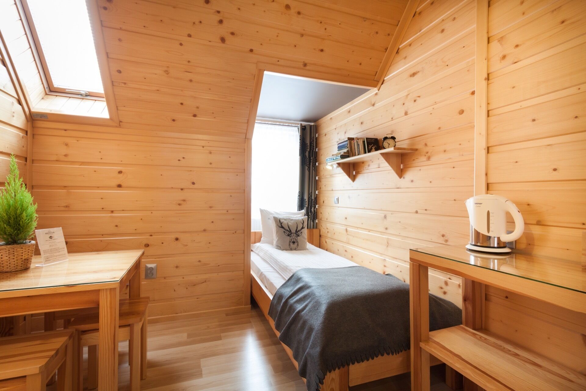 Ein Holzhüttenraum mit einem Bett, einem kleinen Tisch, einem Regal mit Büchern und einem Wasserkocher.