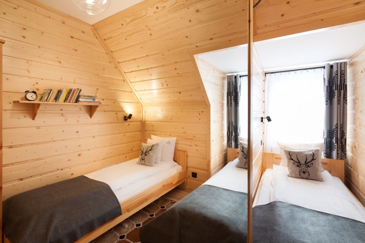 Een hutje met twee houten bedden, een plank met boeken en een klok, en een raam met gordijnen.