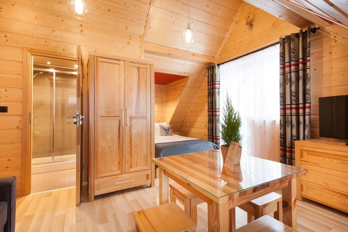 Een knus houten chaletinterieur met een glazen eettafel, een bed met hoofdeinde, een kleine potplant en een televisie op een houten kast.