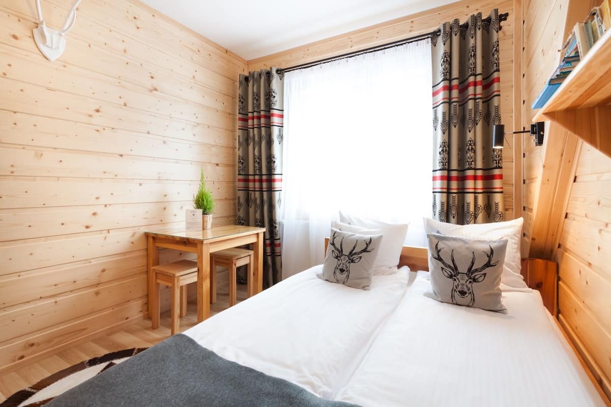 Een houten kamer met een bed, twee kussens met hertenmotieven, een kleine tafel met een plant en ruitgordijnen.