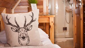 Une chambre avec un plafond en bois, un lit avec un oreiller orné d’une tête de cerf, une petite table avec une plante et une cabine de douche en verre.