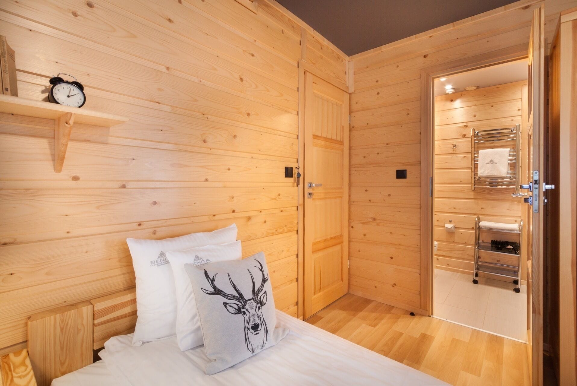 A wooden room with a bed, pillows, a clock, and a bathroom visible through an open door.