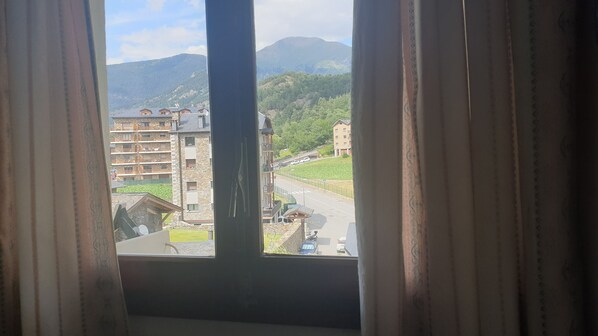 Une vue par une fenêtre, avec des rideaux, donnant sur une rue, des immeubles et une montagne en arrière-plan.