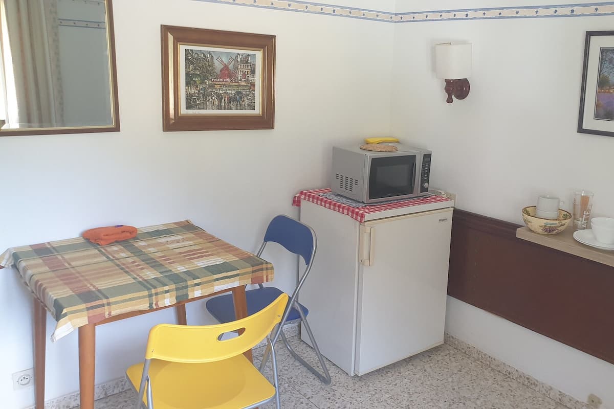 Una cocina compacta con microondas, un refrigerador pequeño, una mesa con mantel a cuadros, una silla amarilla y un estante con cafetera y tazas.