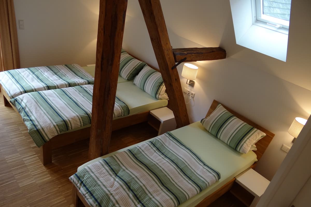 A room with three wooden bunk beds, each with striped bedding. There is a window with a view of the outside, and a wall-mounted lamp on the right side.
