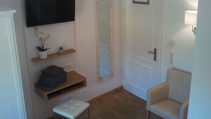 A hotel room with a television mounted on the wall, a mirror, a door, a small white bench, and a chair. - Landgasthaus zum Seysingshof (Heldburger Unterland)