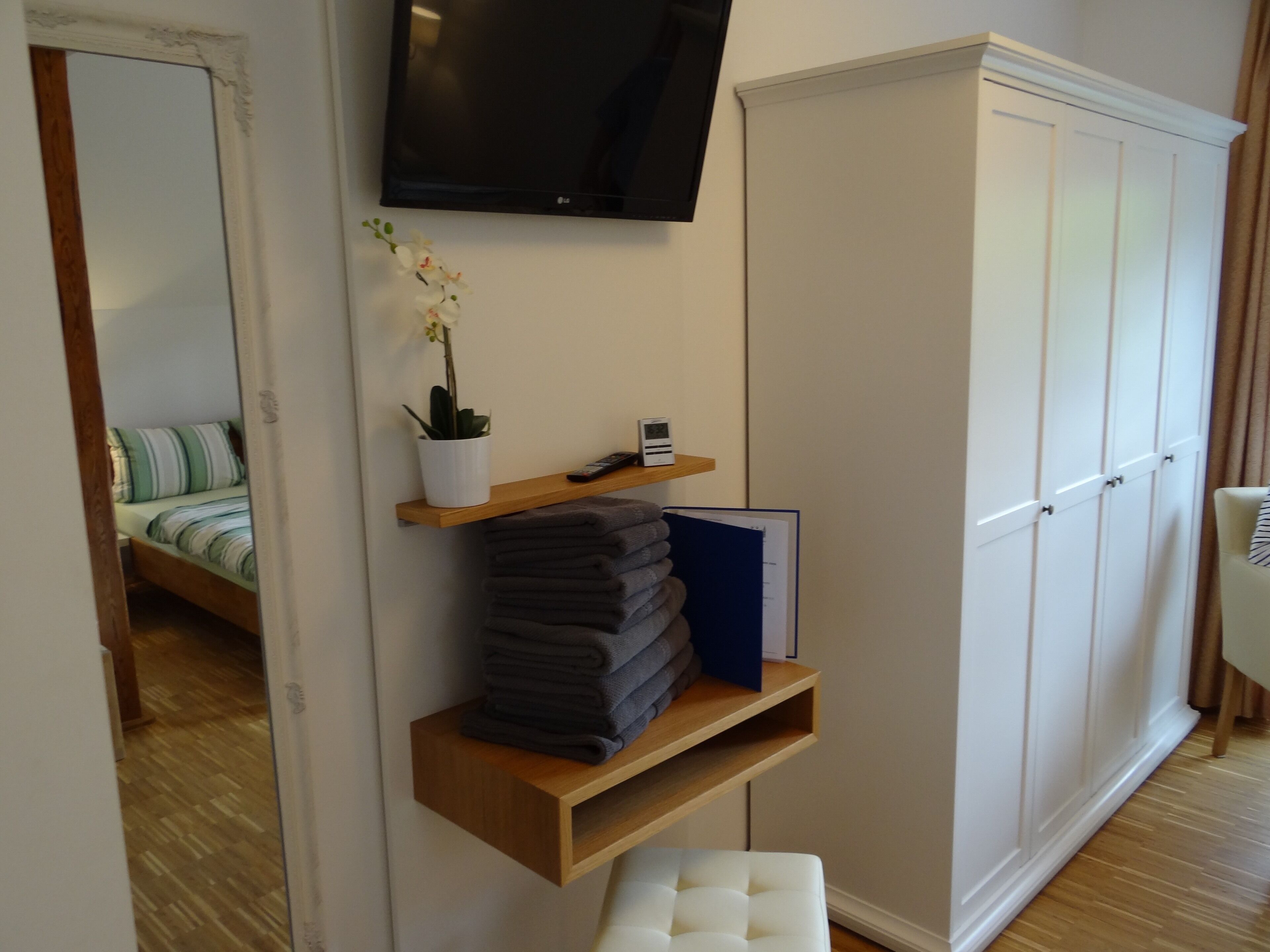 A bedroom with a bed, a TV mounted on the wall, a wardrobe, a shelf with folded clothes, and a potted plant.