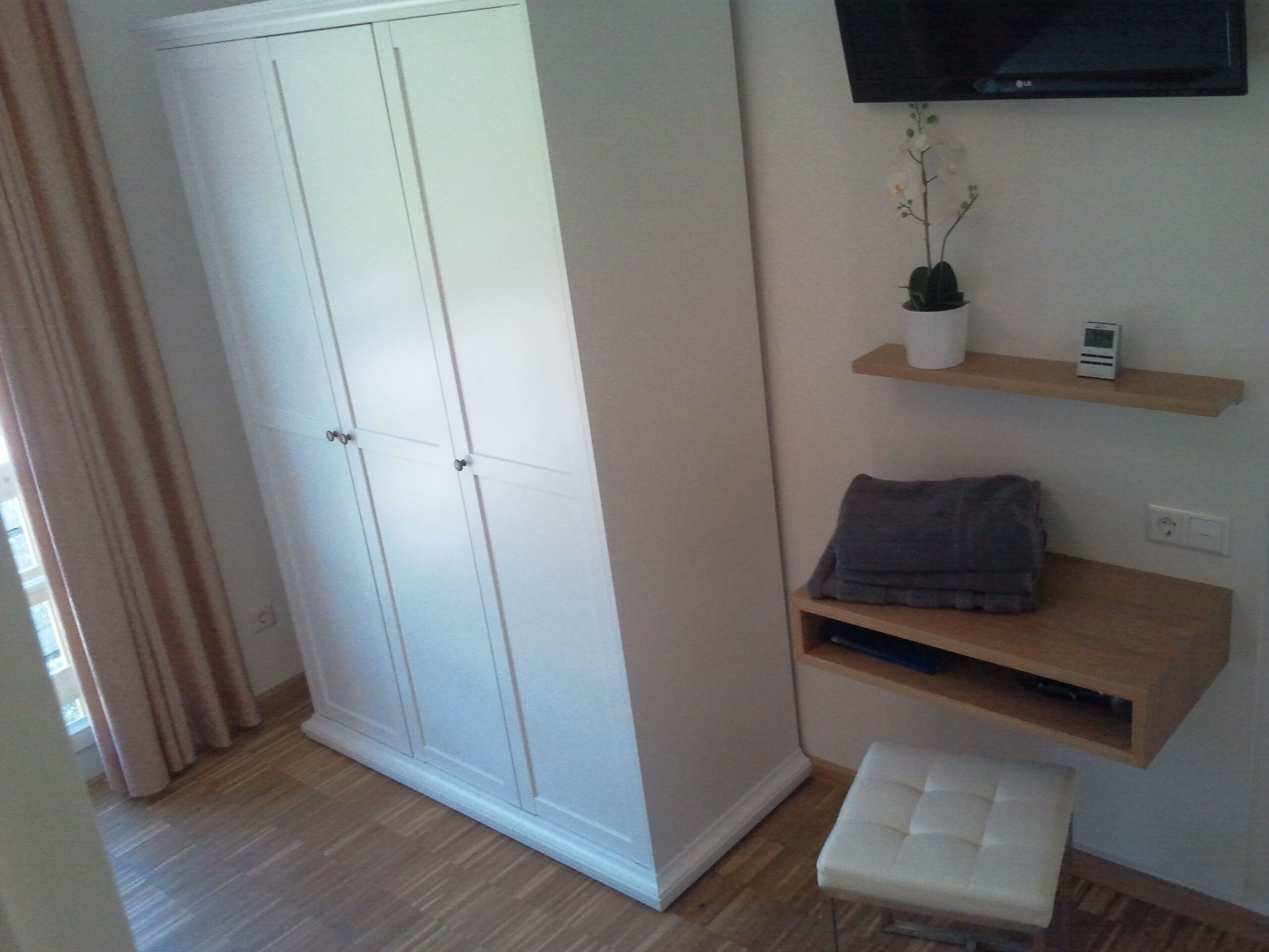 A room with a wardrobe, a TV mounted on the wall, a small shelf with a plant, and a bench.