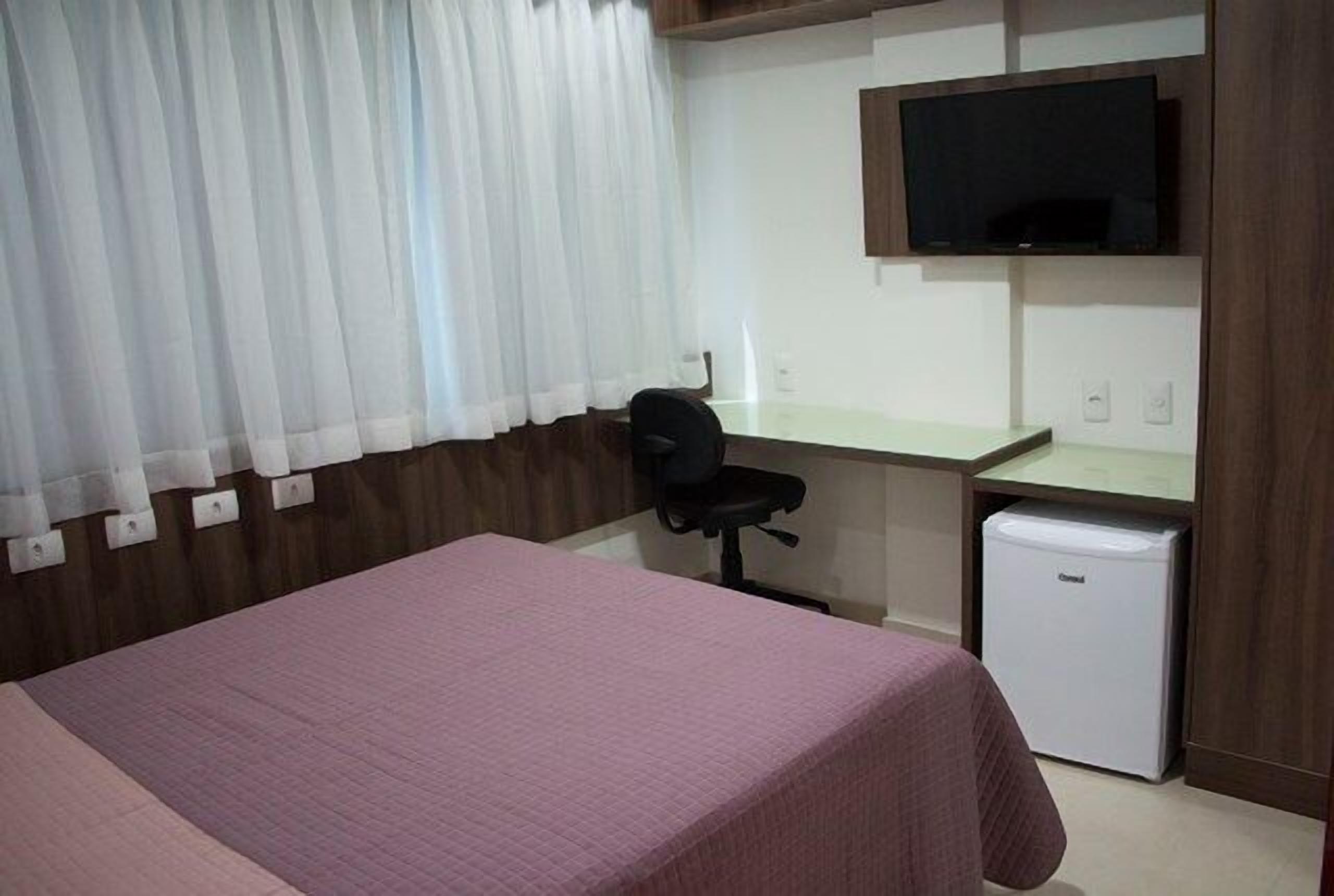 A hotel room with a bed, a desk, a chair, a television, and a small refrigerator.