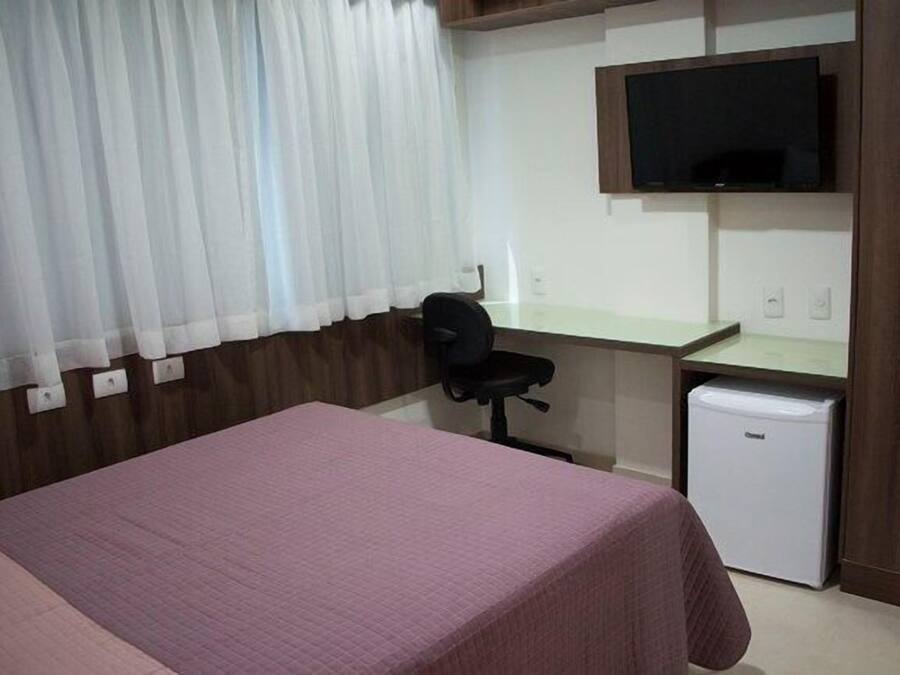A hotel room with a bed, a desk, a chair, a television, and a small refrigerator.
