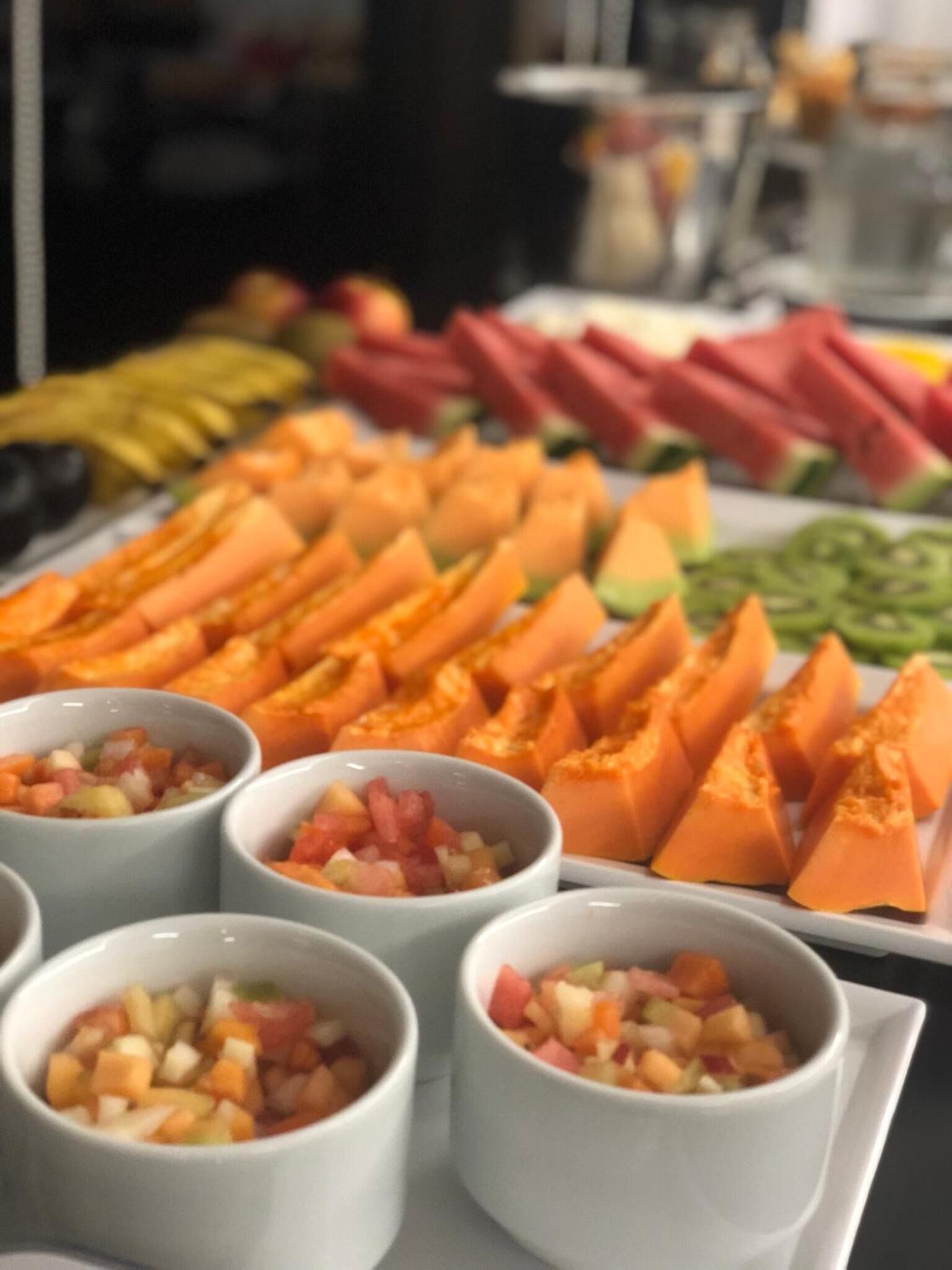 Een buffet met diverse fruit- en groentesgerechten, waaronder gesneden watermeloen, kiwi en wortelen, en kleine schaaltjes gemengde fruitsalade.