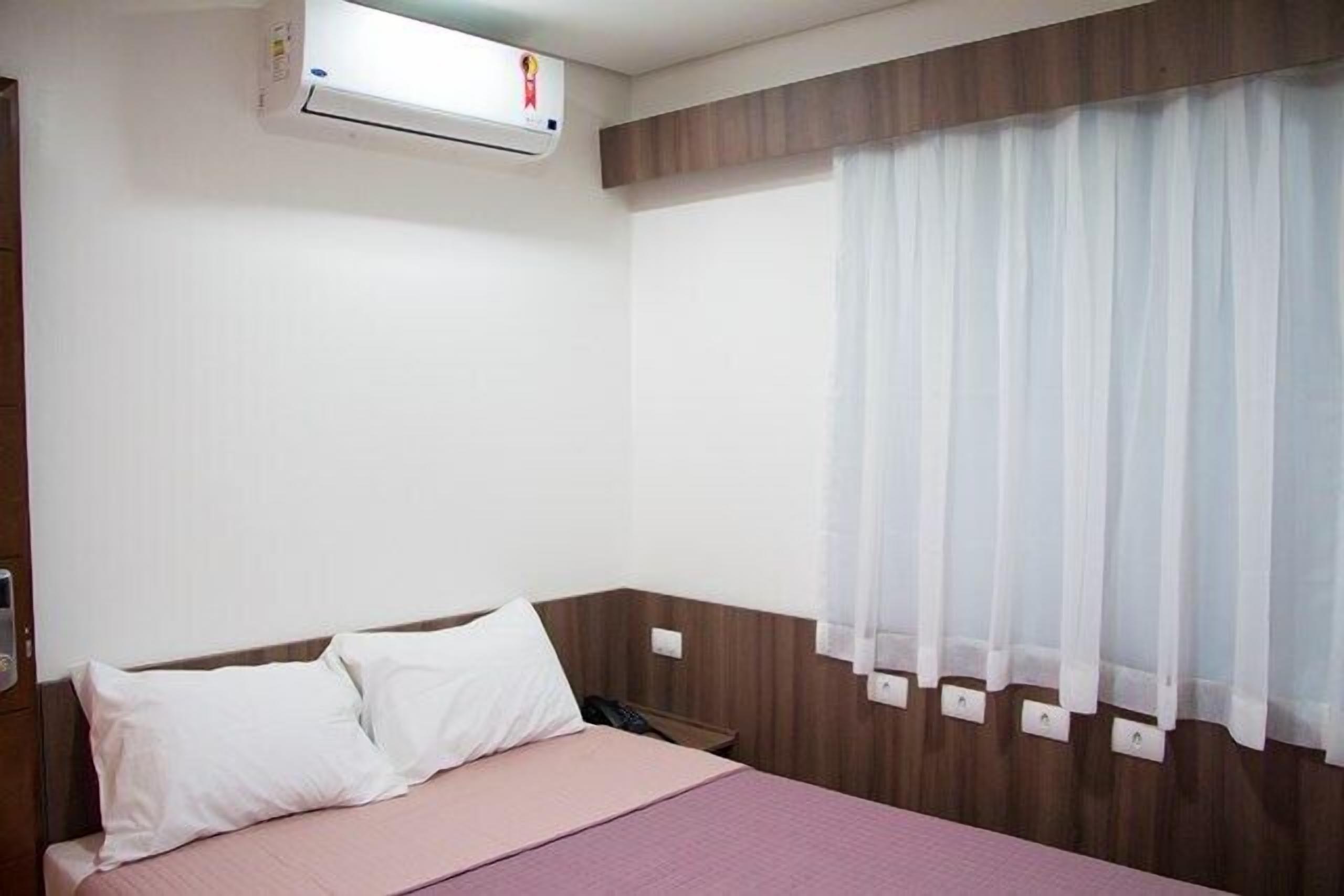 Habitación de hotel con cama, ventana con cortinas translúcidas, unidad de aire acondicionado y cabecero de madera.