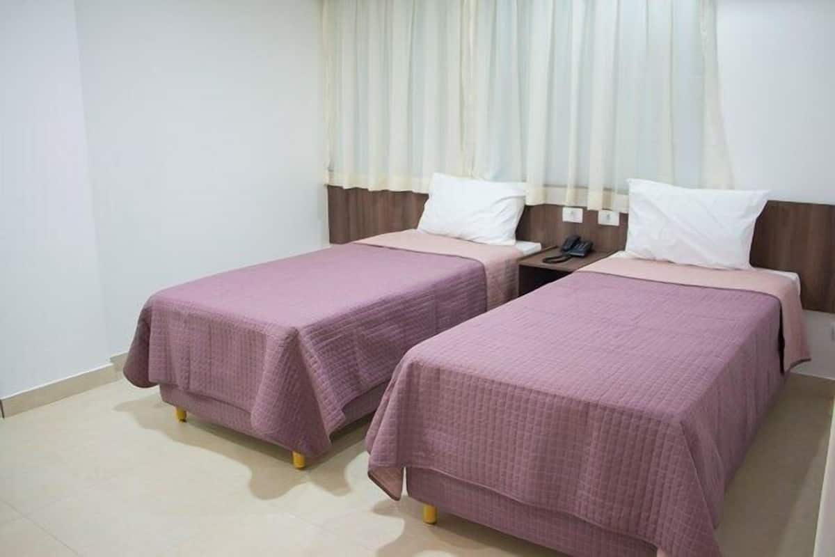 A hotel room with two single beds, each with a pink bedspread and white pillows, a nightstand with a phone, and a window with sheer curtains.