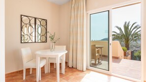 A room with a white table and chairs, a vase with a plant, a large mirror, and a sliding glass door leading to an outdoor area with a view of palm trees and a distant landscape.