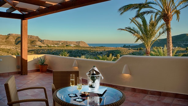 Une terrasse avec une table dressée pour deux personnes, offrant une vue imprenable sur un paysage pittoresque, avec des palmiers et des collines lointaines.
