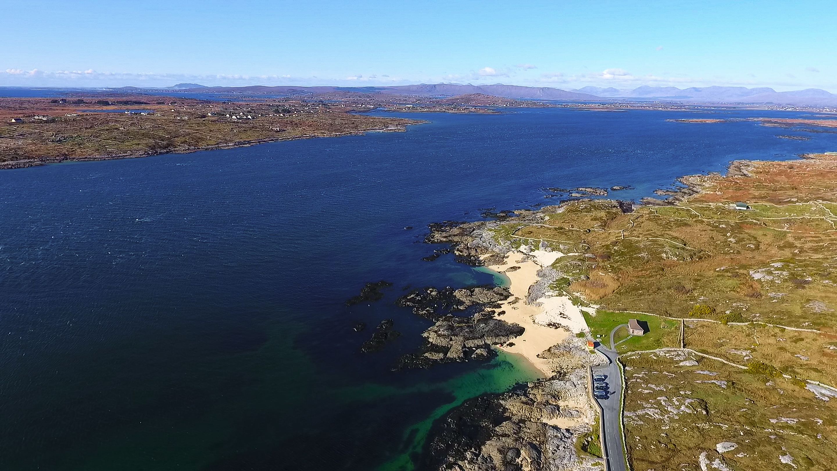 An aerial view of a coastal area with a road leading to a beach, surrounded by rocky terrain and sparse vegetation.