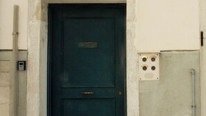 A dark blue door with a small window above it, a doorbell, and a utility box with four switches on the wall. - Mario Apartment 5012 (Venice)