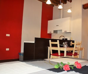 A room with a dining table set for two, a kitchen area with white cabinets, and a red accent wall.