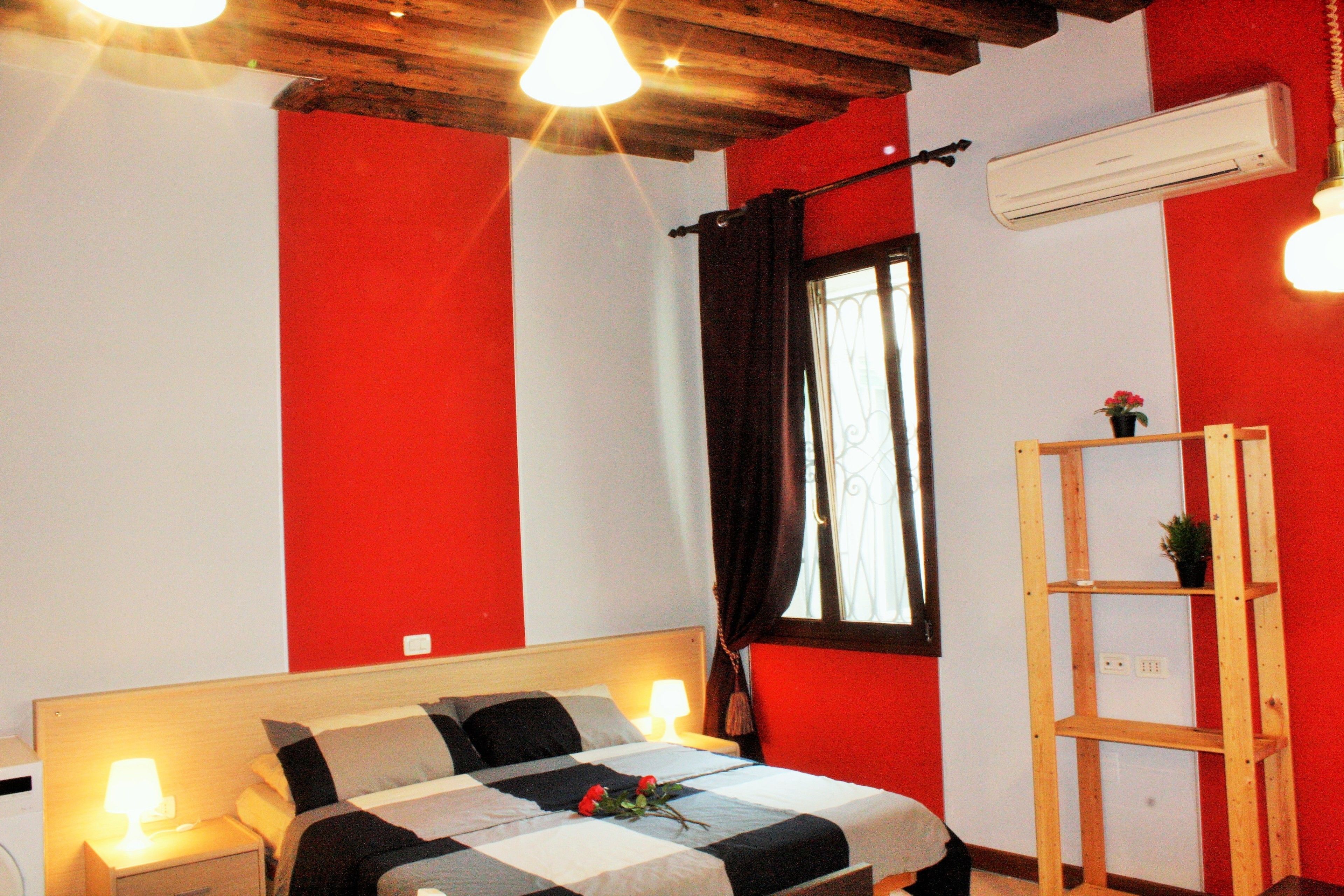 A bedroom with a bed, bedside lamps, a window with curtains, an air conditioning unit, and a wooden shelf.