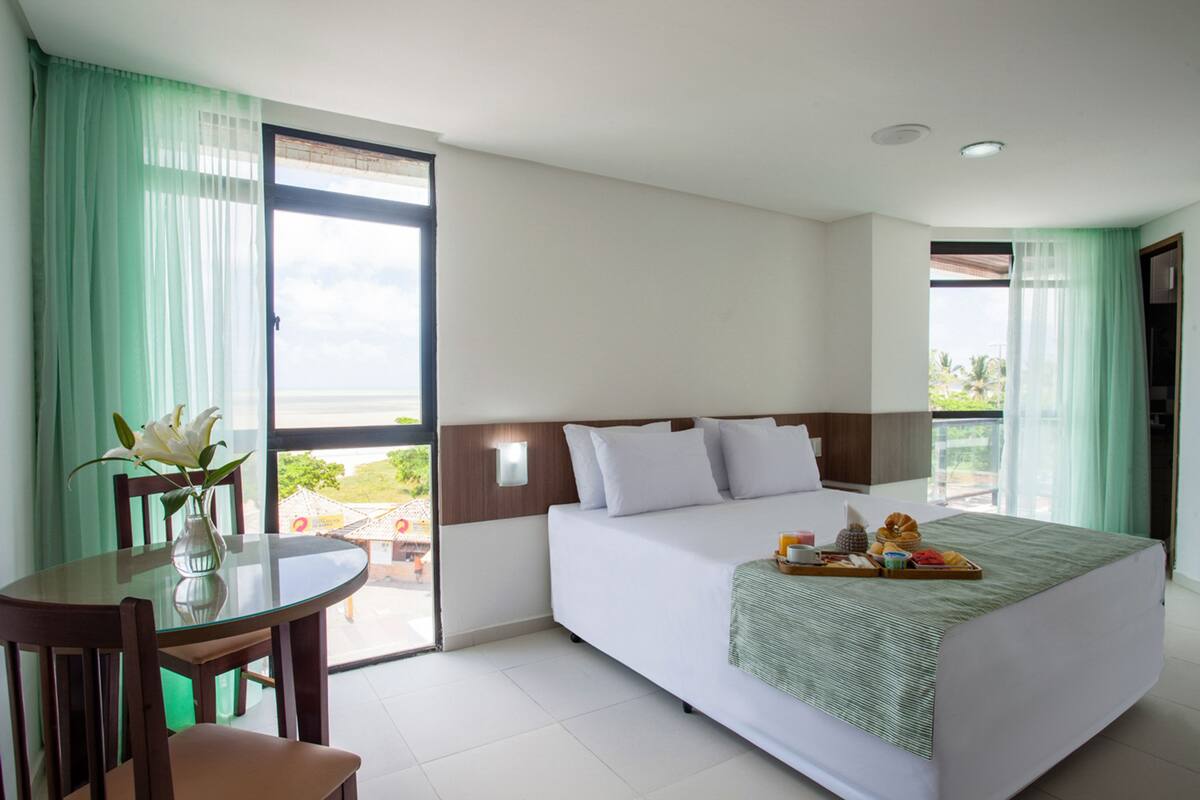 Habitación de hotel con cama, una mesa de comedor con sillas, un jarrón con flores y un balcón con vistas.