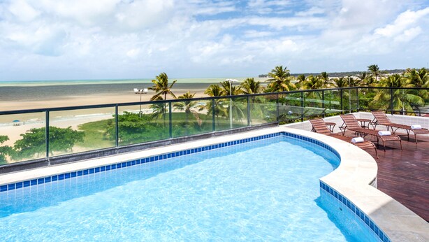 Una piscina con borde curvo, barandilla de vidrio y vista a la playa y palmeras.