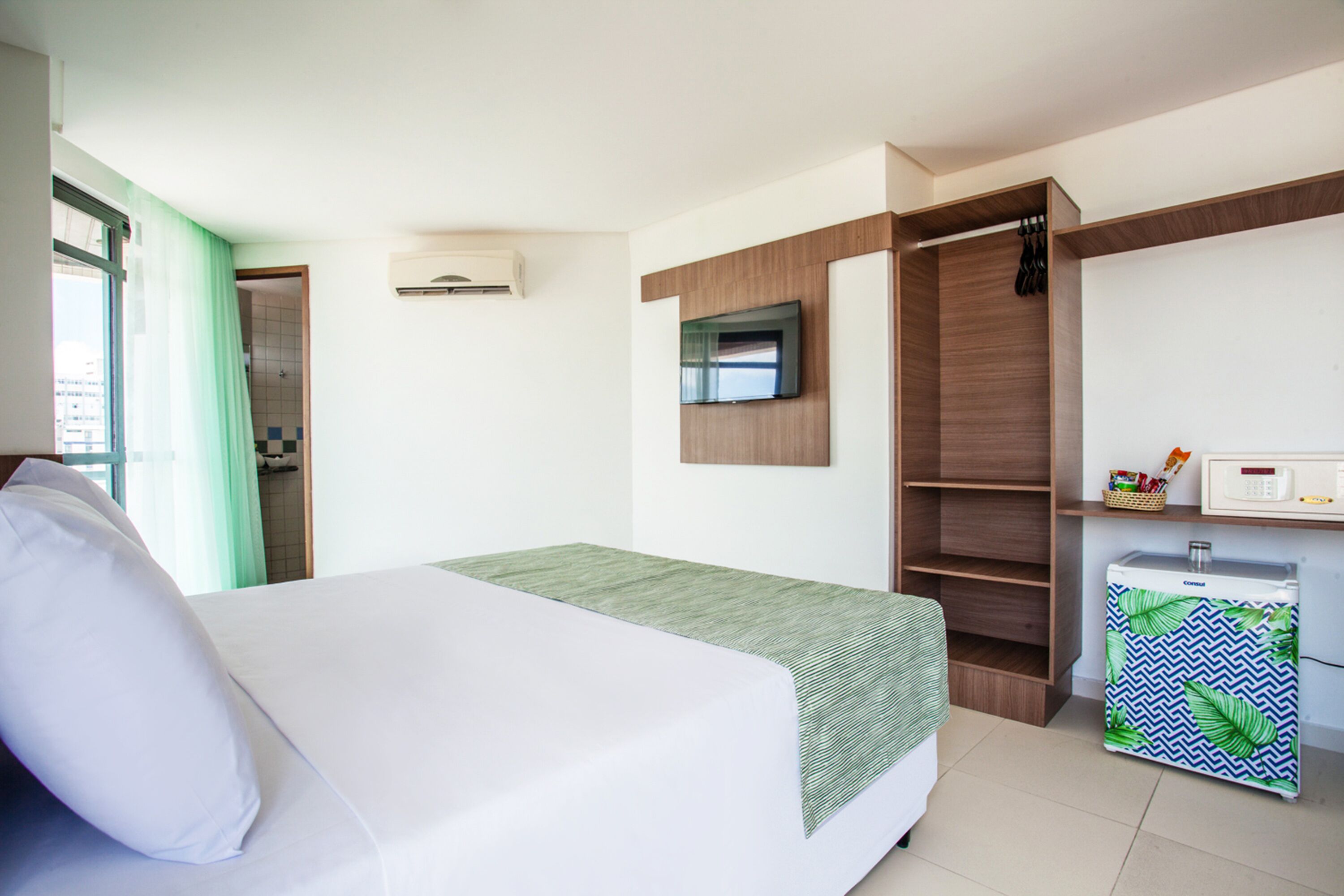 Habitación de hotel con cama, televisor montado en la pared, armario y mini refrigerador.