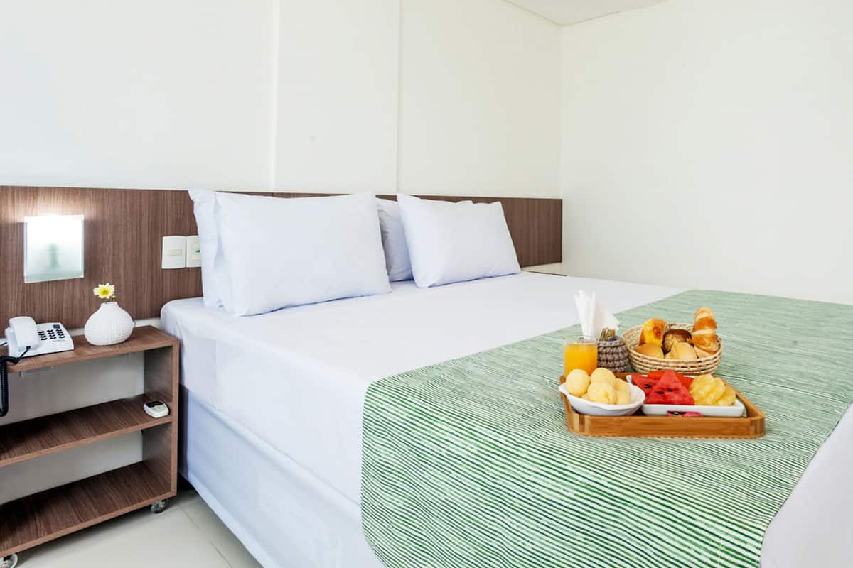 Habitación de hotel con una cama, una mesita de noche con un teléfono y un florero, y una bandeja de desayuno con frutas y bollería.