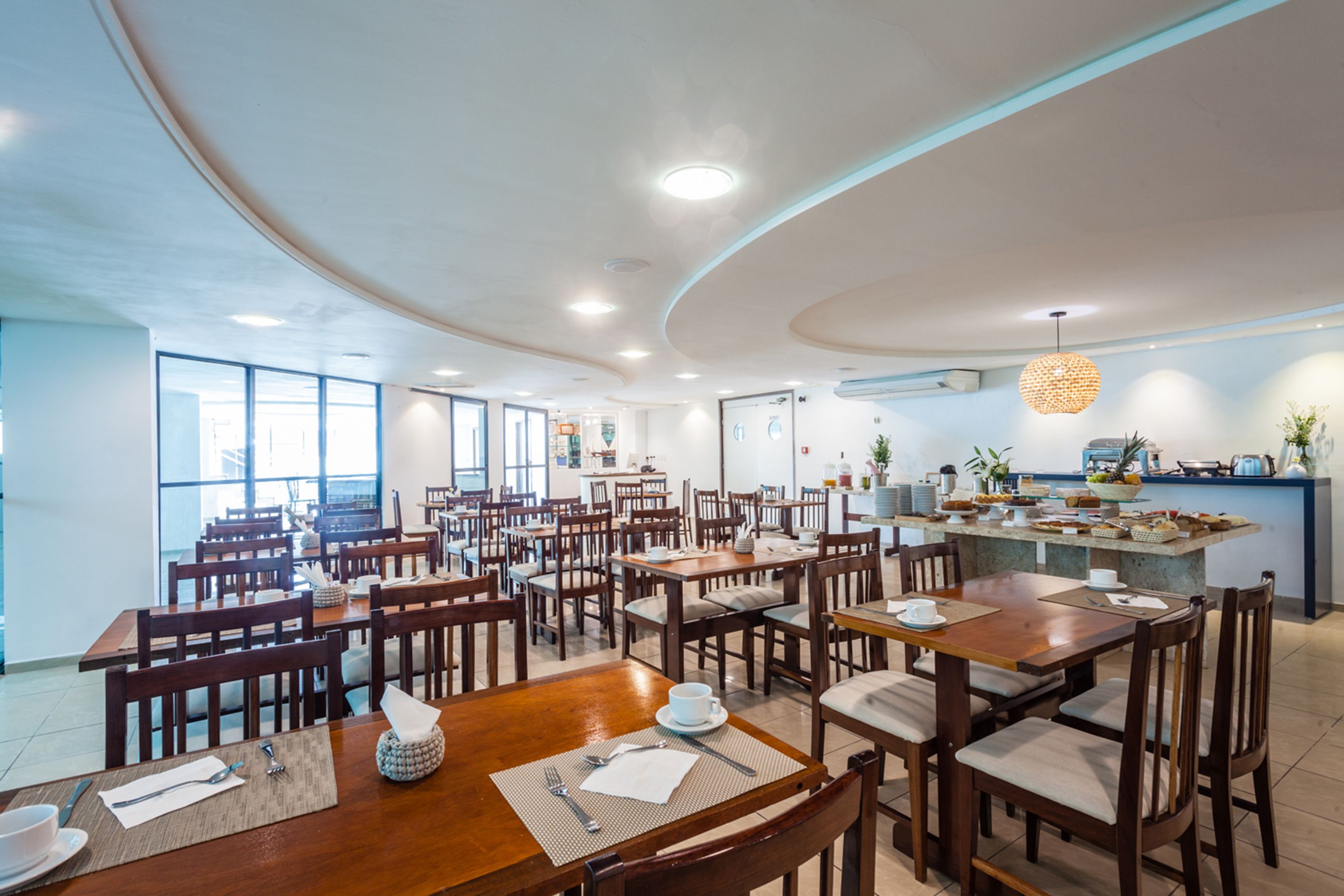 Un comedor amplio con varias mesas y sillas de madera, una isla central con encimera de cocina y ventanas grandes que dejan entrar luz natural.