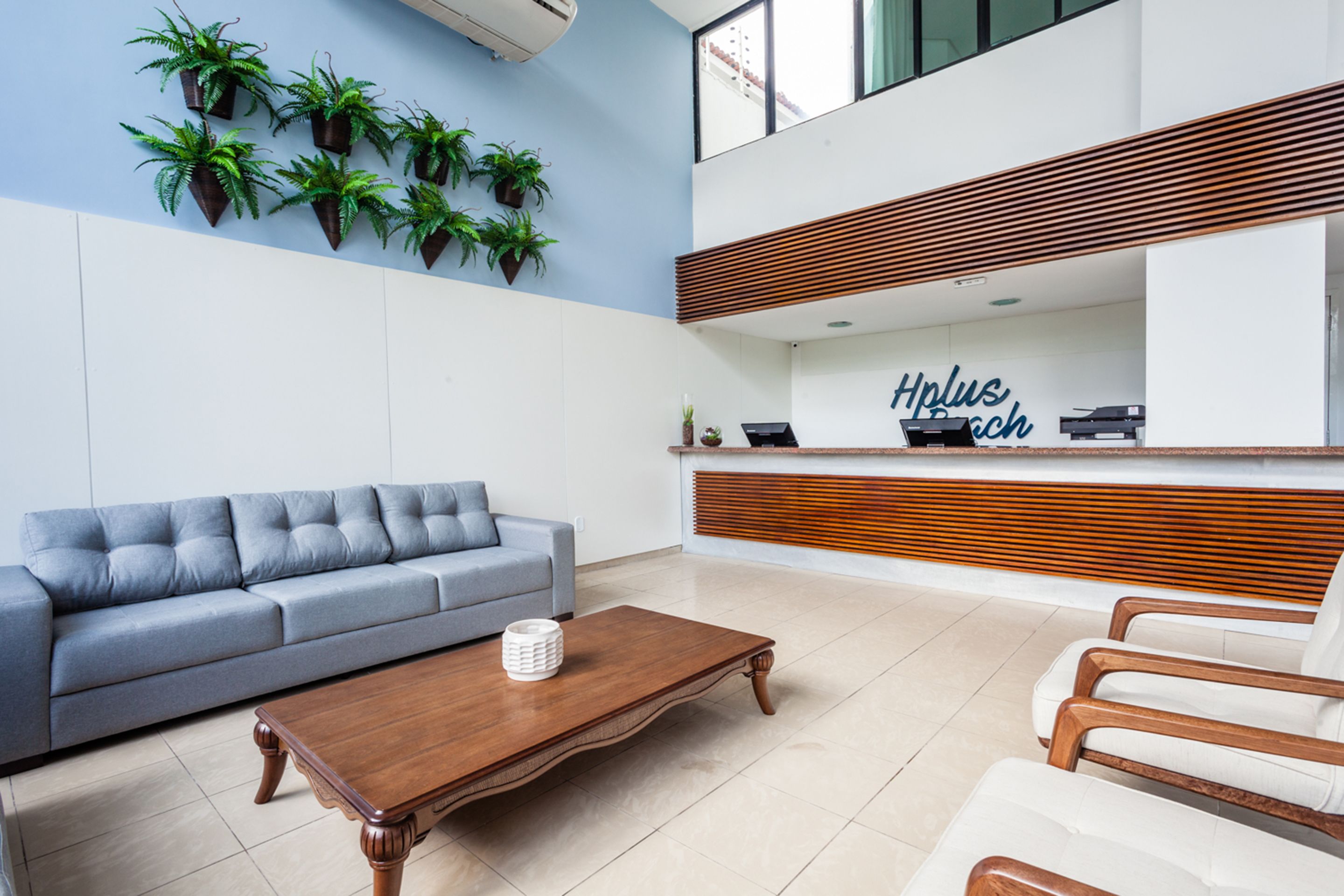 Un área de recepción moderna con un sofá gris, una mesa de centro de madera y un mostrador de recepción con un letrero que dice "Hplus beach".