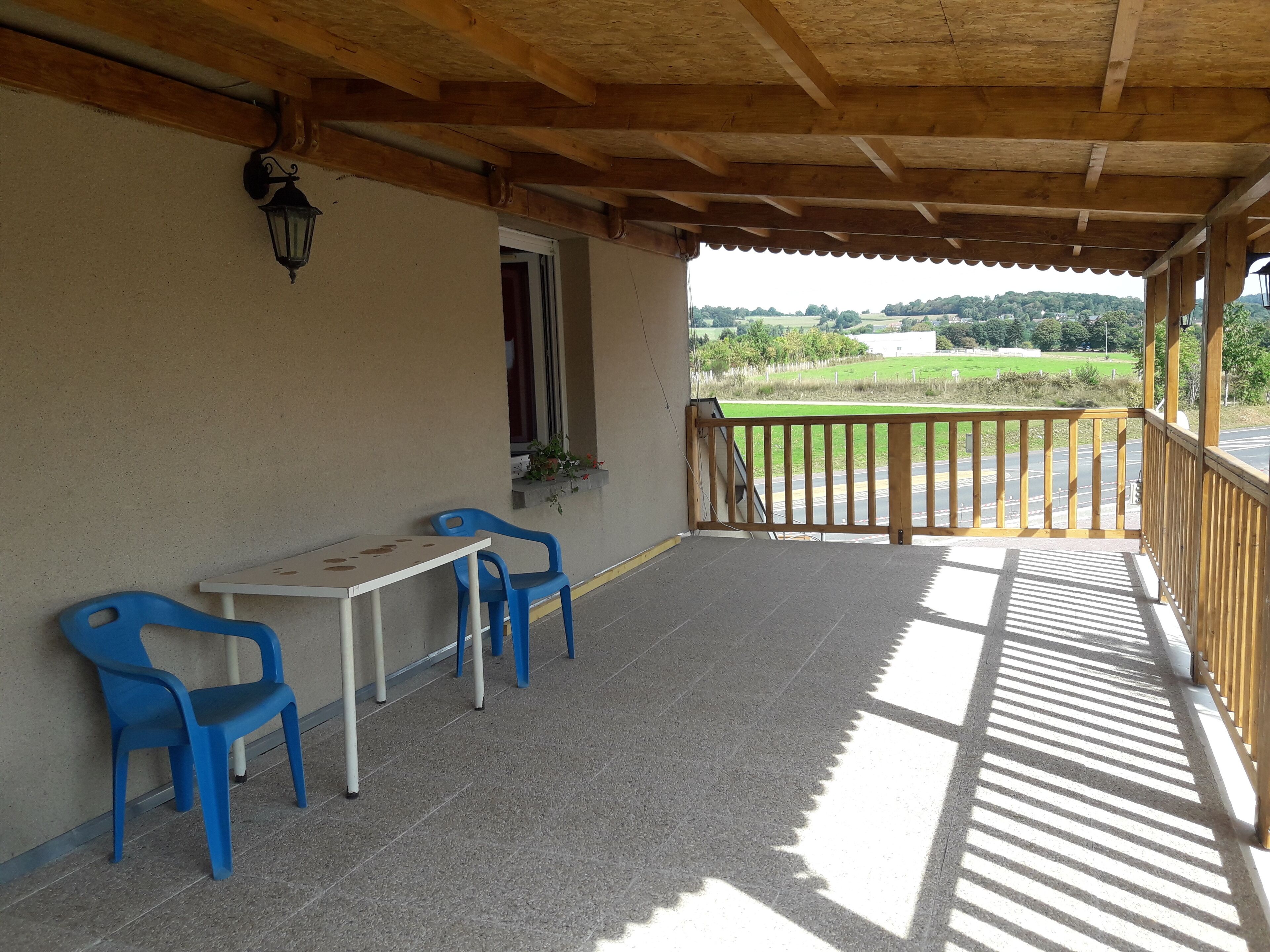A covered outdoor seating area with two blue plastic chairs and a small table, a wooden railing, and a view of a green landscape.