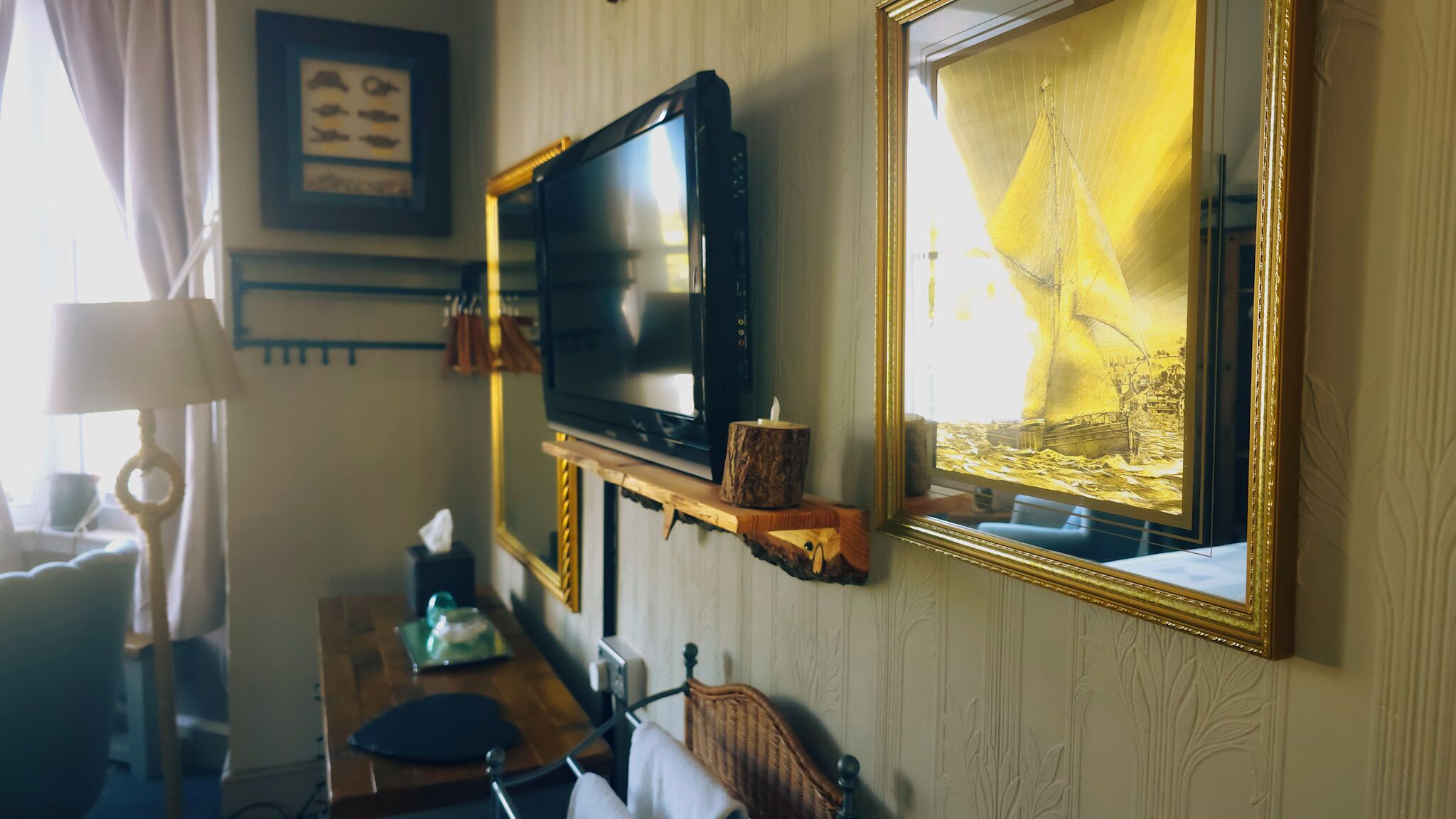 A room with a television mounted on a wooden shelf, a framed picture on the wall, a desk with a chair, and a window with curtains.