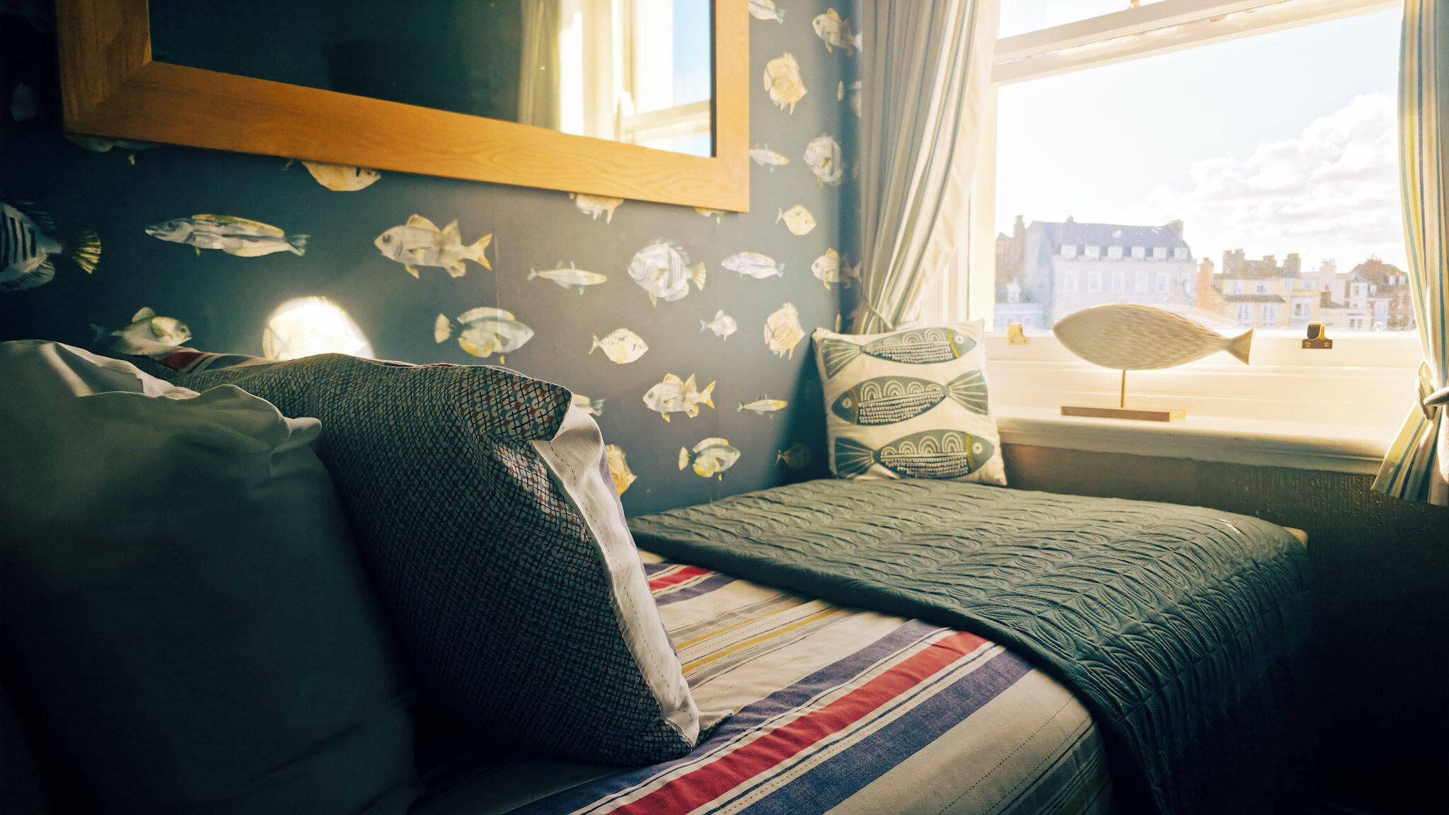 A room with a bed, pillows, a window with a view of buildings, and a wall with fish patterns.
