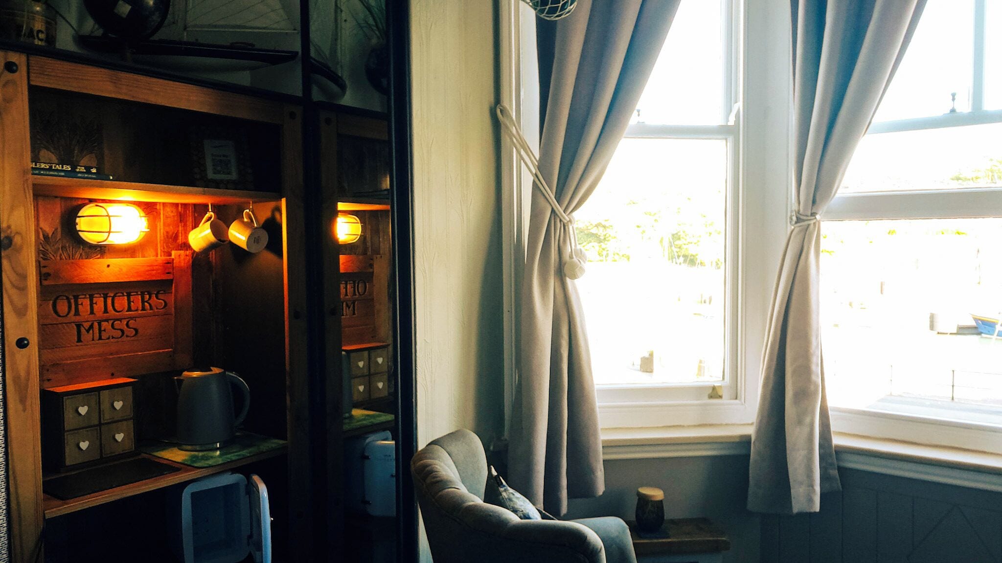 Ein Zimmer mit einem Holzbodenregal, auf dem 'OFFICERS MESS' steht, einem Stuhl, einem Fenster mit Vorhängen und Blick nach draußen.