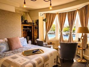 Quarto de hotel com cama, poltrona, escrivaninha, abajur e vista para a água.
