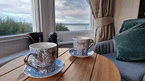 Two patterned cups on saucers on a wooden table by a window with a view of a body of water and greenery. - The Ocean Guesthouse (Weymouth)